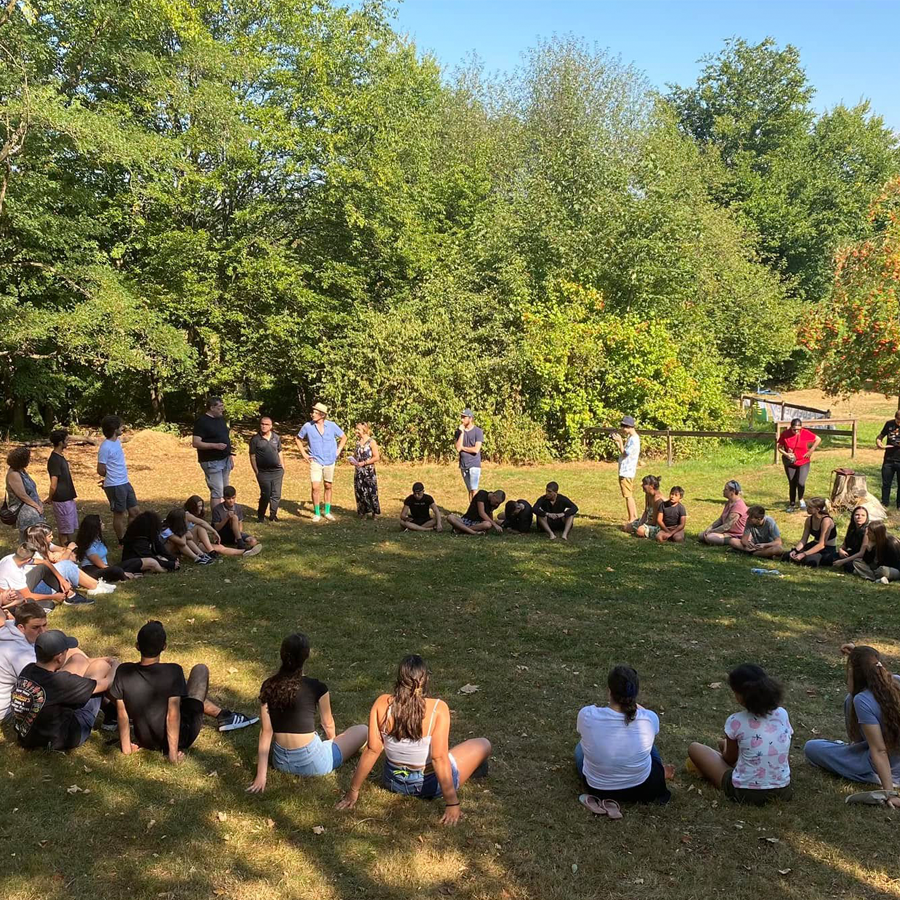 Jugendliche aus Deutschland, Tschechien und Israel sitzen während eines trilateralen Jugendaustauschs im sächsischen Bahratal in einem großen Kreis auf einer Wiese. Die Kennenlernrunde findet unter freiem Himmel statt.